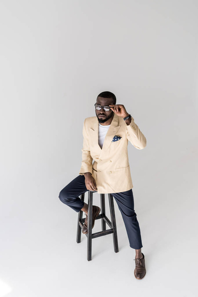 high angle view of handsome young african american man adjusting eyeglasses and sitting on stool isolated on grey - Foto, afbeelding