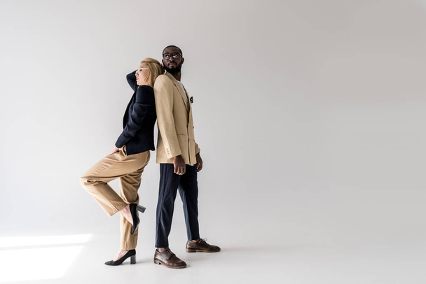 side view of fashionable young multiethnic couple posing back to back on grey - Foto, imagen