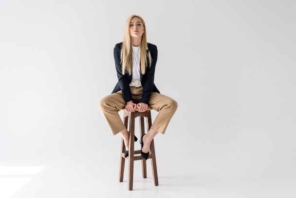 beautiful young blonde woman in stylish clothes sitting on stool and looking at camera isolated on grey  - Photo, Image