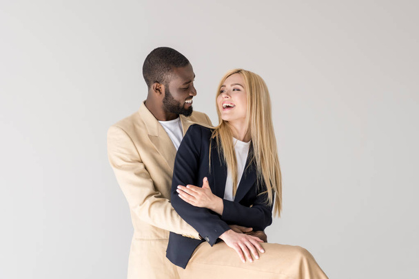 happy stylish young multiethnic couple hugging and smiling each other isolated on grey - Foto, Imagem