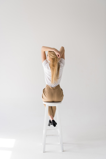 Widok z młoda kobieta siedzi na krześle i dotykając blond włosy na szary tyłu  - Zdjęcie, obraz
