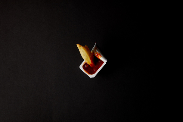 κάτοψη του πλαστικού εμπορευματοκιβωτίου με κέτσαπ με χύνεται τηγανιτής πατάτας που απομονώνονται σε μαύρο - Φωτογραφία, εικόνα