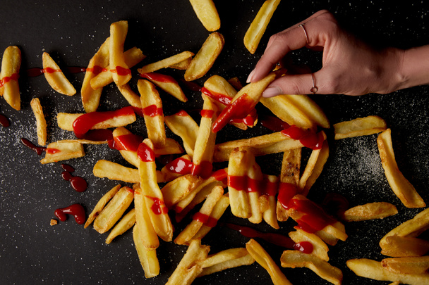 top view of woman taking french fries poured with ketchup on black tabletop - Photo, Image