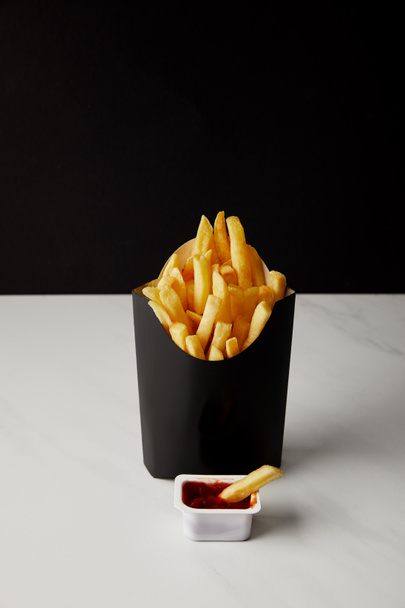 box of french fries with plastic container of ketchup on white marble surface isolated on black - Фото, изображение