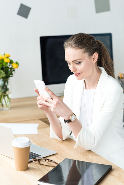 femme d'affaires souriante en costume élégant en utilisant smartphone sur le lieu de travail dans le bureau
 - Photo, image