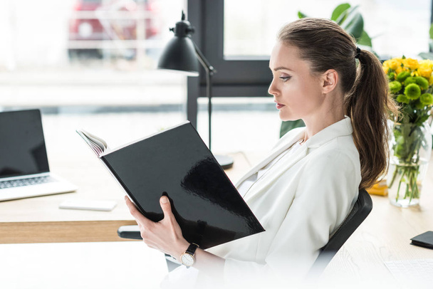 side view of beautiful businesswoman with book at workplace in office - Foto, immagini
