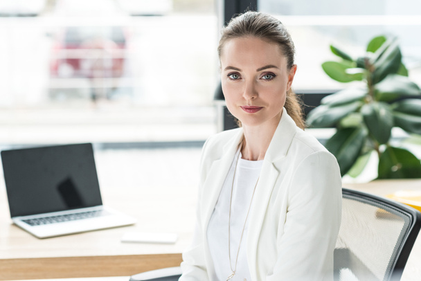 portrait of smiling beautiful businesswoman in white suit at workplace with laptop in office - Photo, Image