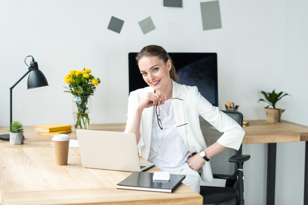 sorridente bela mulher de negócios em terno branco sentado no local de trabalho com laptop e café para ir no escritório
 - Foto, Imagem