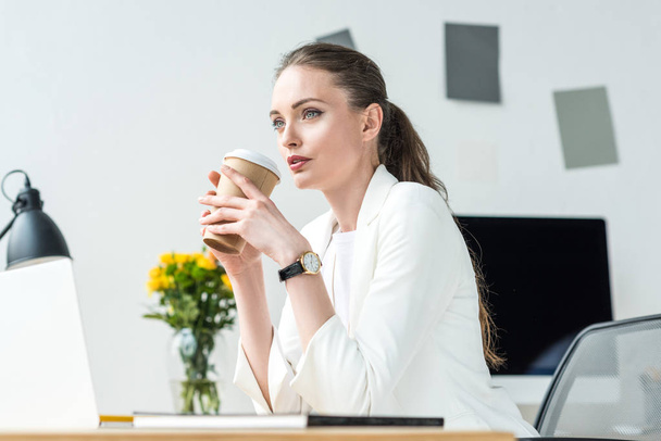 Zijaanzicht van peinzende zakenvrouw met koffie te gaan op de werkplek op kantoor - Foto, afbeelding