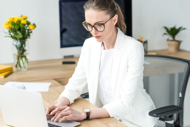 focused businesswoman in eyeglasses working on laptop at workplace in office - Photo, Image