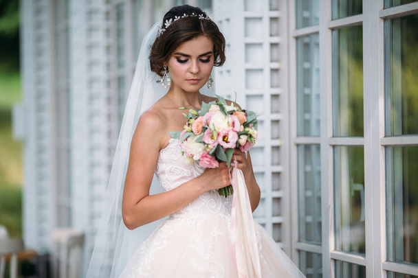 Portrait of beautiful bride with bouquet flowers in white luxury dress. Gorgeous model with bridal makeup and hairstyle with a diadem in her hair, diamond earrings in the ear. Marriage and floristics - Foto, afbeelding