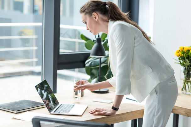 vista laterale di donna d'affari con vetro d'acqua utilizzando il computer portatile sul posto di lavoro in ufficio
 - Foto, immagini
