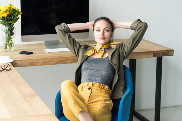 beautiful stylish businesswoman sitting with hands behind head at workplace and looking at camera  - Фото, изображение