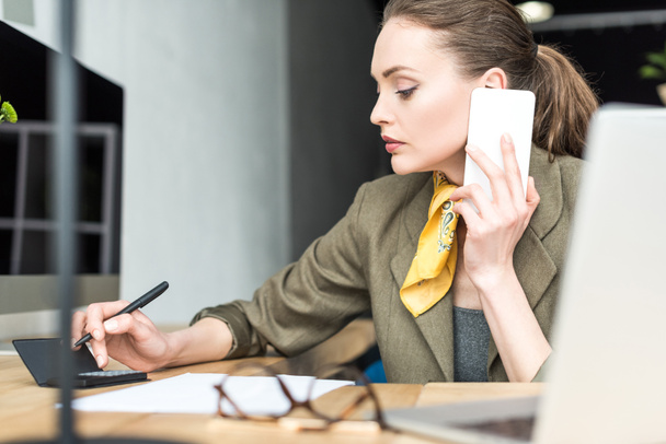 concentrated businesswoman using calculator and talking by smartphone in office - Photo, Image