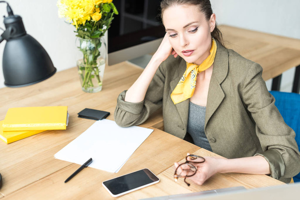 high angle view of beautiful stylish businesswoman holding eyeglasses and looking away while sitting at workplace - Photo, Image