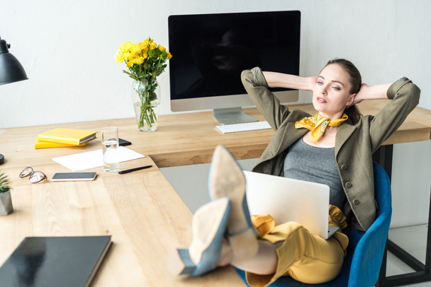 κομψό επιχειρηματίας με laptop κάθεται με τα χέρια πίσω από το κεφάλι και βλέπει μακριά στο γραφείο - Φωτογραφία, εικόνα