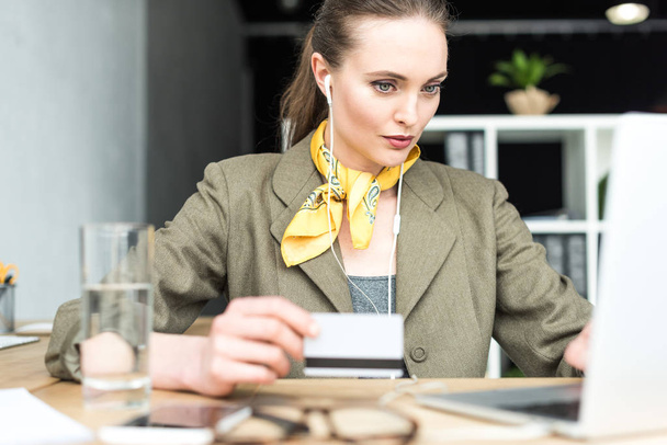 femme d'affaires élégante dans les écouteurs tenant la carte de crédit et en utilisant un ordinateur portable au bureau
 - Photo, image
