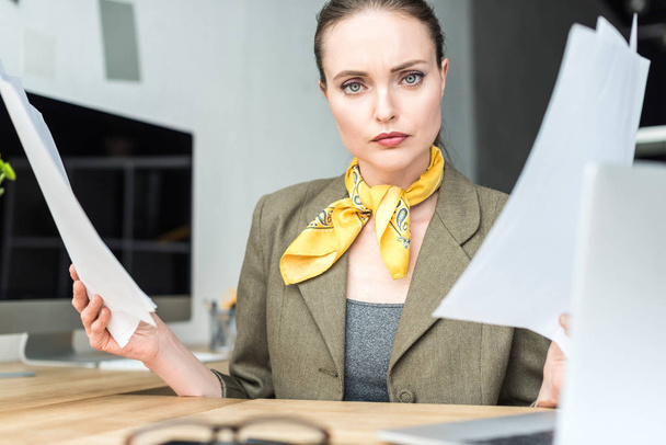 seria mujer de negocios con estilo sosteniendo papeles y mirando a la cámara en la oficina
 - Foto, Imagen