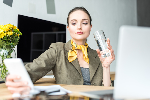 attractive stylish businesswoman drinking water and using smartphone at workplace - Zdjęcie, obraz