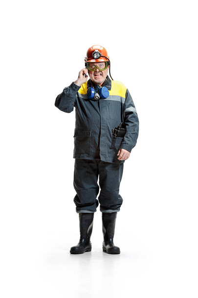 The studio shot of angry senior bearded male miner standing at the camera with smartphone on a white background. - Foto, afbeelding
