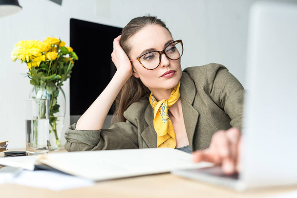 attractive stylish businesswoman in eyeglasses using laptop at workplace - Fotoğraf, Görsel