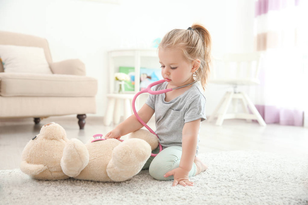 Cute little girl with stethoscope and toy bear playing at home - 写真・画像