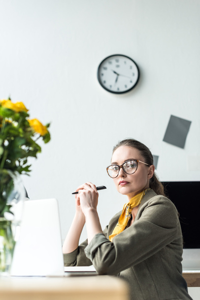 beautiful middle aged businesswoman in eyeglasses using laptop and looking at camera in office - Photo, Image