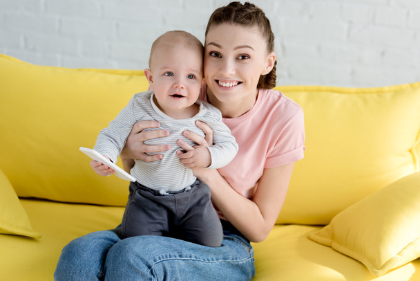 happy mother with little son holding smartphone and sitting on sofa - Foto, Bild
