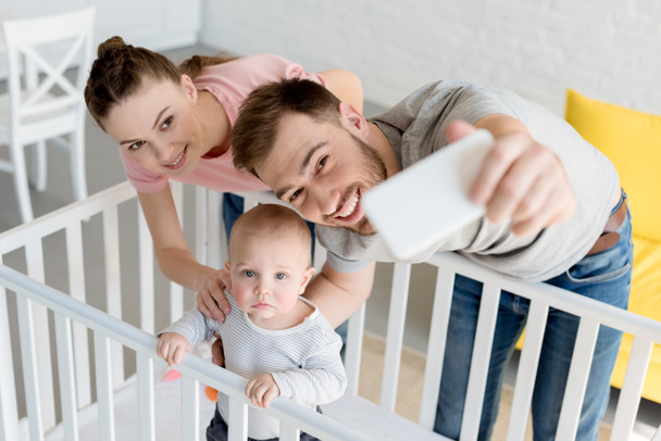 padres sonrientes tomando selfie con su hijo en la cuna en el teléfono inteligente
 - Foto, Imagen