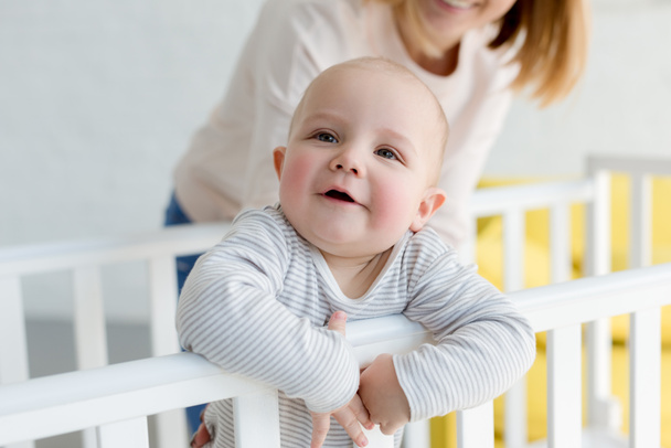 pequeño hijo sonriente en cuna de bebé con madre detrás
 - Foto, Imagen