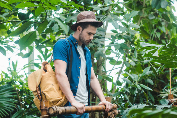 thoughtful young man in stylish clothes with backpack in park with lot of tropical plants - Photo, image