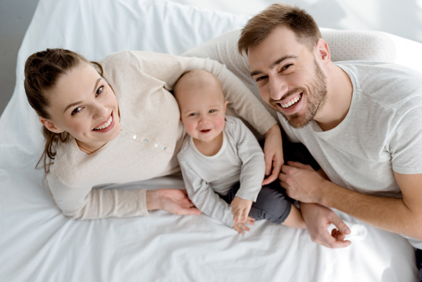 bovenaanzicht van gelukkige jonge ouders met baby liggend op bed - Foto, afbeelding
