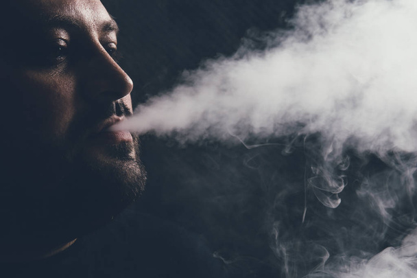 Człowieku vape e papierosa z e ciecz, zbliżenie, oddycha się obłoku pary lub pary na ciemnym tle. Koncepcja vaping - Zdjęcie, obraz