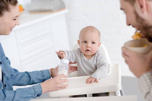 parents feeding baby boy with milk in baby bottle on kitchen - Fotó, kép