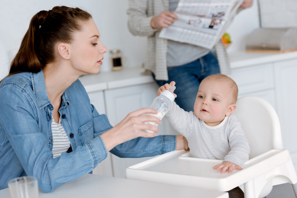 Äiti ruokkii pientä poikaa maitoa tuttipullossa keittiössä, isä sanomalehden takana
 - Valokuva, kuva