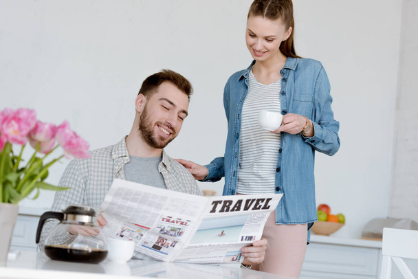 husband reading travel newspaper while smiling wife with coffee standing near in kitchen - Photo, Image