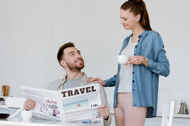 husband reading travel newspaper while wife with coffee standing near - Фото, изображение