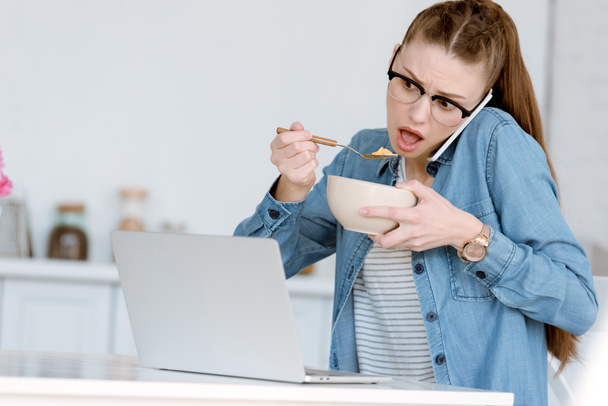 σοκαρισμένος γυναίκα teleworker τρώει πρωινό ενώ μιλώντας στο smartphone και ψάχνει για φορητό υπολογιστή  - Φωτογραφία, εικόνα