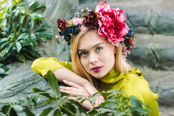 beautiful young woman with floral wreath leaning on rocks and looking at camera - Photo, Image