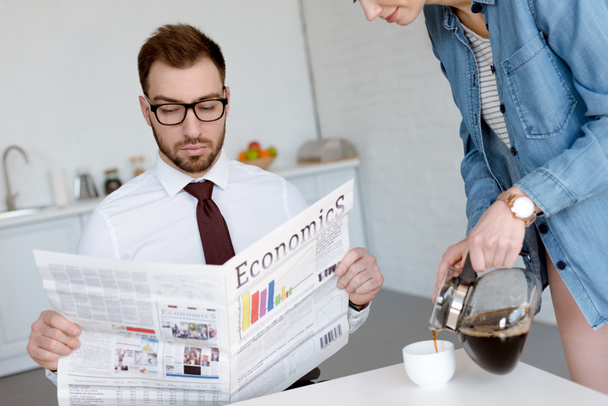 hombre de negocios en anteojos leyendo el periódico de economía mientras esposa vertiendo café en taza en la cocina
 - Foto, Imagen