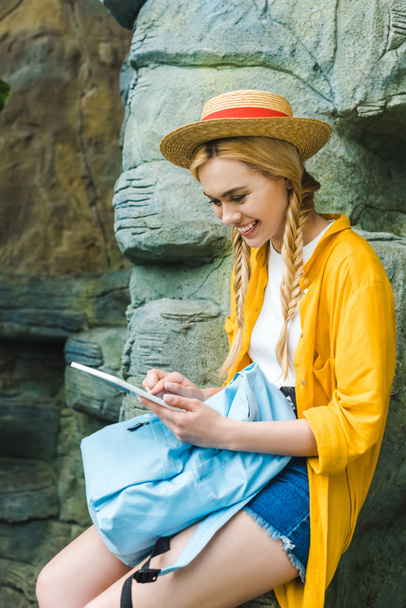 smiling young woman in straw hat using tablet while sitting on rocks outdoors - Photo, Image