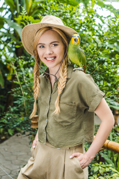 Lächelnde junge Frau im Safarianzug mit Papagei auf der Schulter im Dschungelpark - Foto, Bild