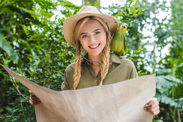 attraktive junge Frau im Safarianzug mit Papagei auf der Schulter navigiert im Dschungel mit Karte - Foto, Bild