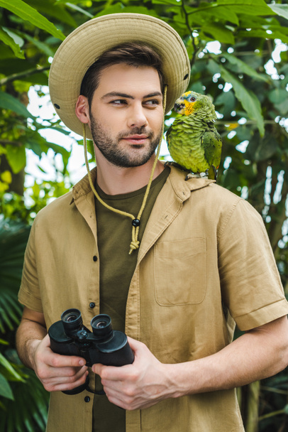 schöner junger Mann im Safarianzug schaut Papagei auf Schulter im Dschungel an - Foto, Bild