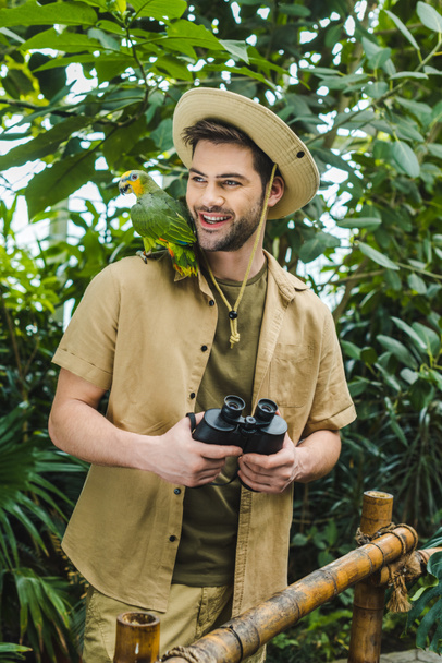 smiling young man with parrot on shoulder and binoculars in jungle - Photo, Image