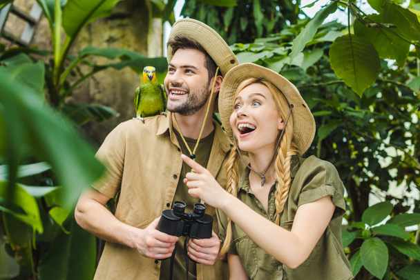 excited young couple in safari suits with parrot in rainforest - Foto, Imagen