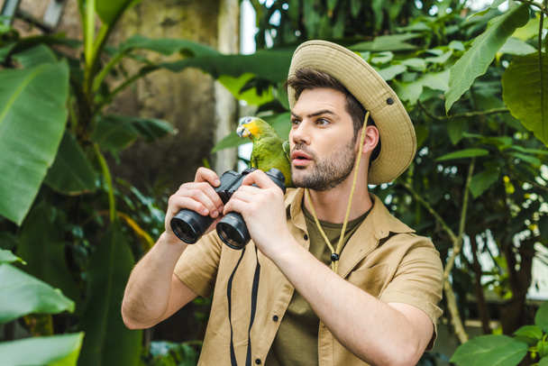 shocked young man with parrot on shoulder and binoculars in jungle - Photo, Image