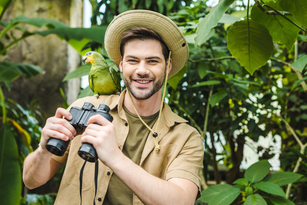 Gelukkig jongeman met papegaai op schouder en verrekijker camera kijken - Foto, afbeelding