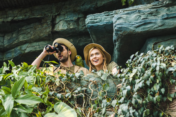 bottom view of excited young couple in safari suits with binoculars on cliff in jungle - Photo, Image