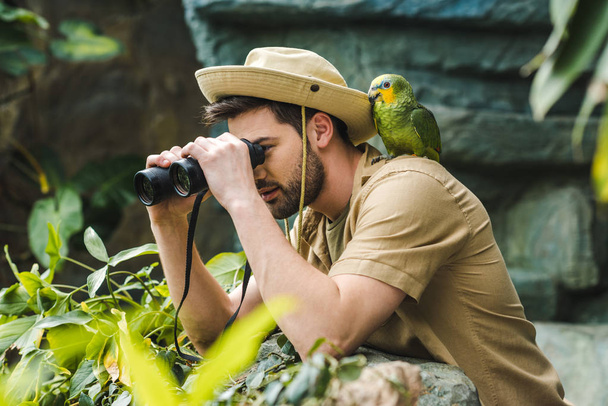 handsome young man with parrot on shoulder looking through binoculars in jungle - Photo, Image